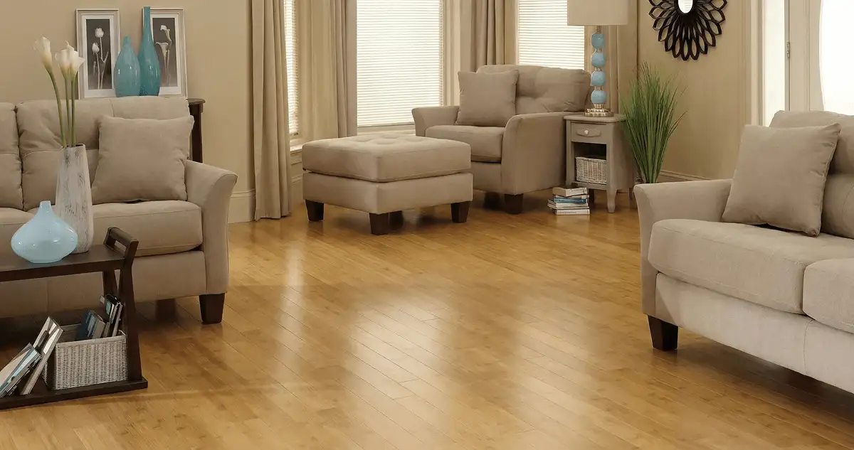 A living room with professionally installed bamboo flooring, beige sofas, a matching ottoman, a side table with a blue lamp, vases, framed pictures, and large windows with blinds and curtains in Ogden, UT